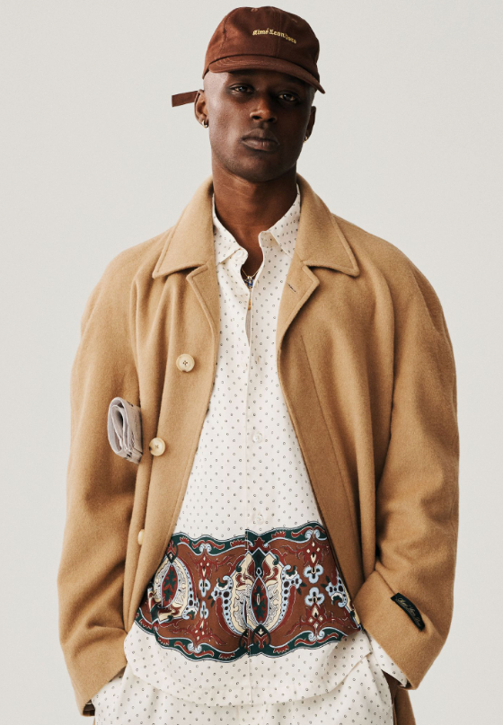 A man wearing a brown Aime Leon Dore trench coat and white button up with a dark brown hat