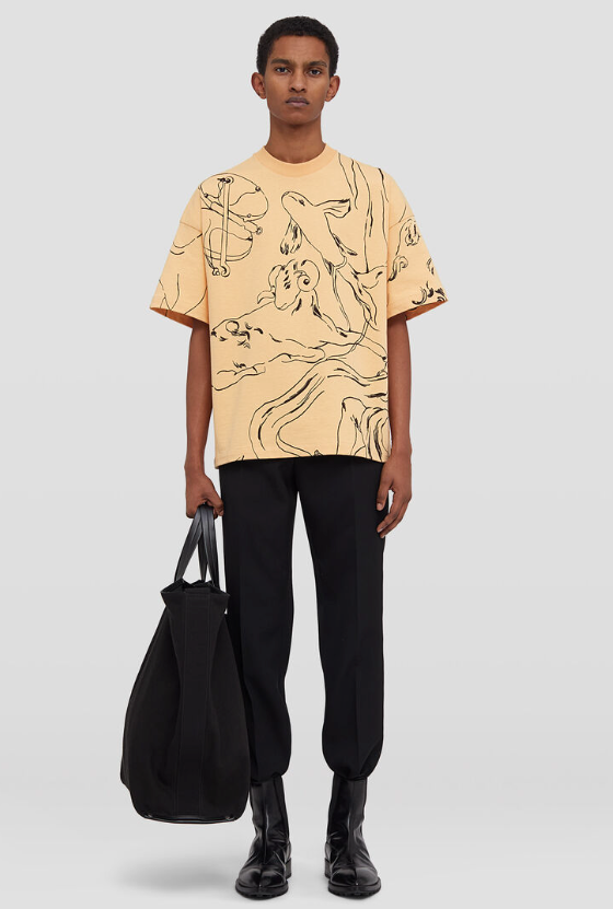 A man wearing a peach colored Jil Sander illustrated short-sleeved t-shirt with black pants and black boots