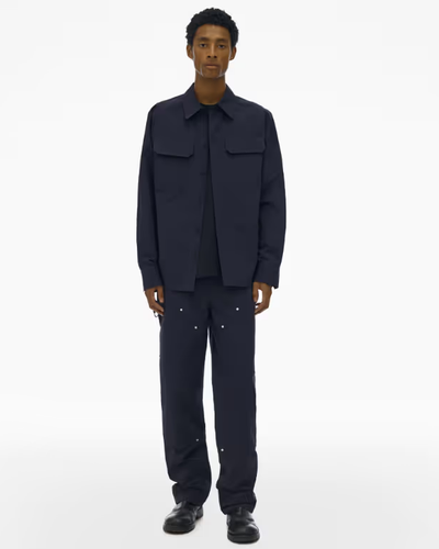 A man wearing a full Navy blue Helmut Lang outfit with a button up top and wide pants and dark navy blue loafers