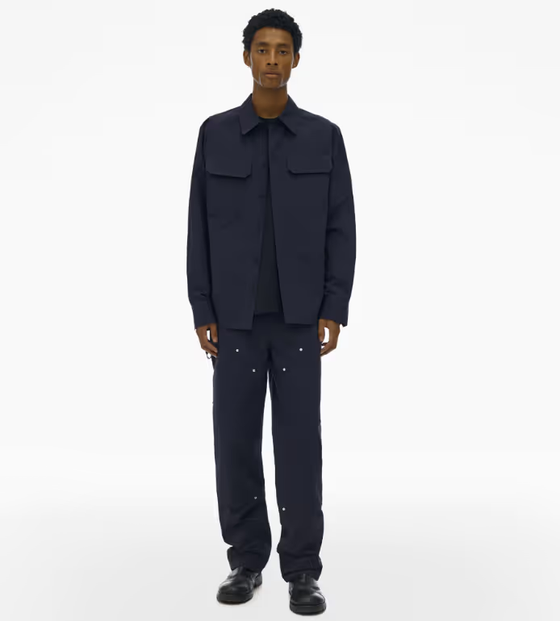 A man wearing a full Navy blue Helmut Lang outfit with a button up top and wide pants and dark navy blue loafers