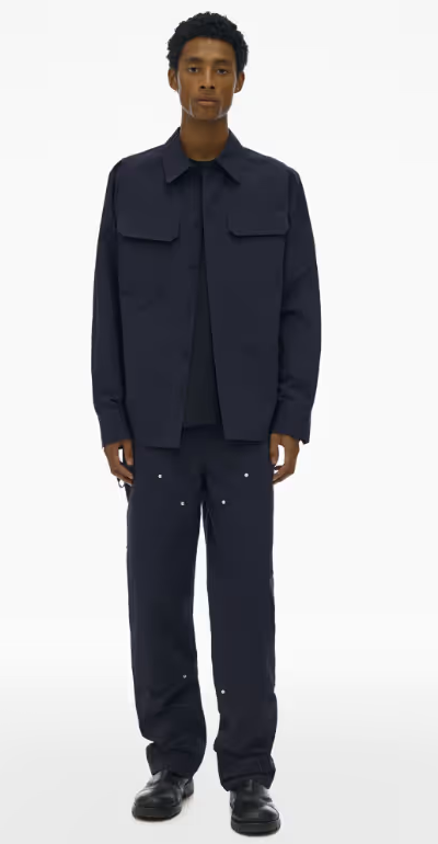 A man wearing a full Navy blue Helmut Lang outfit with a button up top and wide pants and dark navy blue loafers