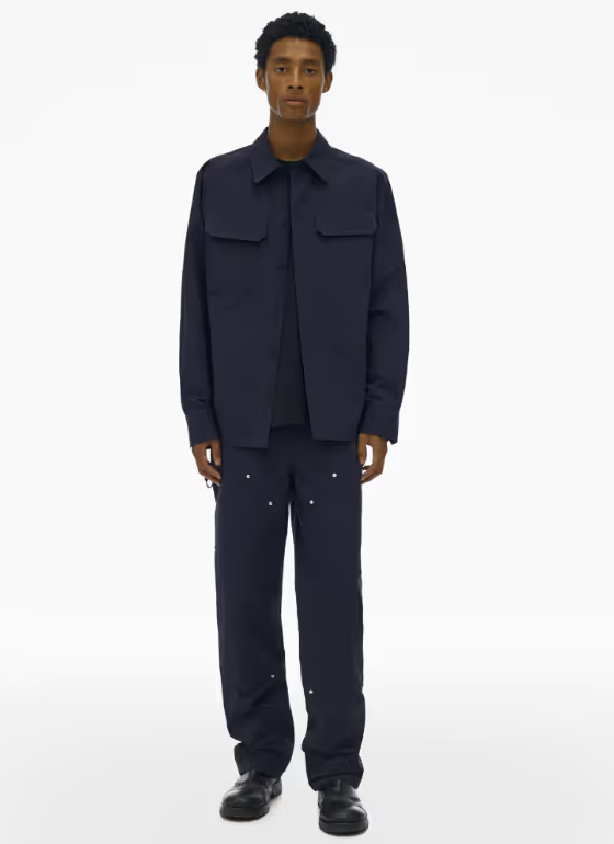 A man wearing a full Navy blue Helmut Lang outfit with a button up top and wide pants and dark navy blue loafers