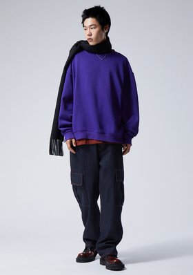 A person wearing a Weekday bright purple sweater, navy blue scarf, denim pants and red and black loafers