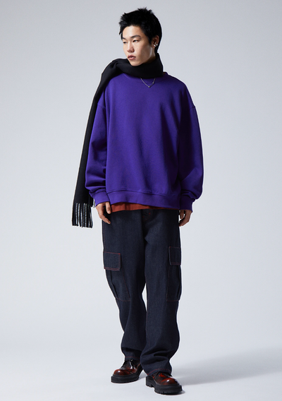 A person wearing a Weekday bright purple sweater, navy blue scarf, denim pants and red and black loafers