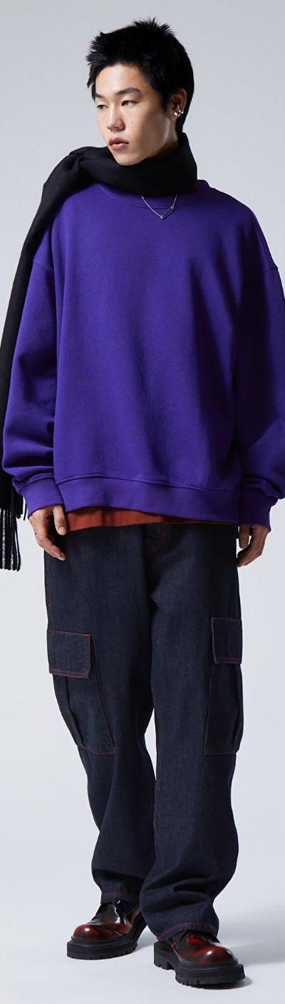 A person wearing a Weekday bright purple sweater, navy blue scarf, denim pants and red and black loafers