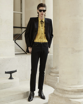 A slim man wearing a Yves St Laurent yellow striped button up with a bolo tie, black blazer, pants and shoes.