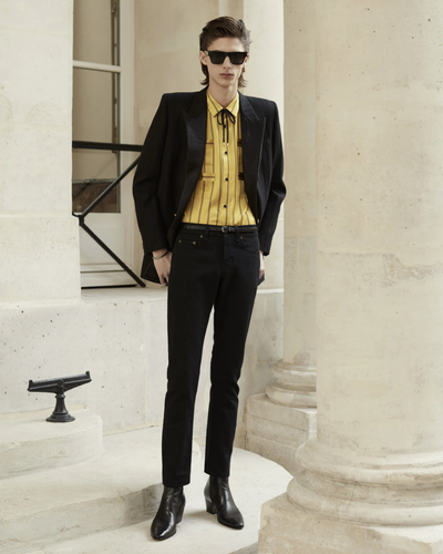 A slim man wearing a Yves St Laurent yellow striped button up with a bolo tie, black blazer, pants and shoes.