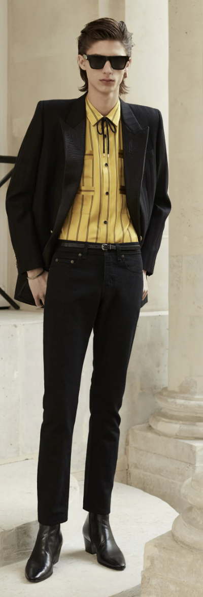 A slim man wearing a Yves St Laurent yellow striped button up with a bolo tie, black blazer, pants and shoes.