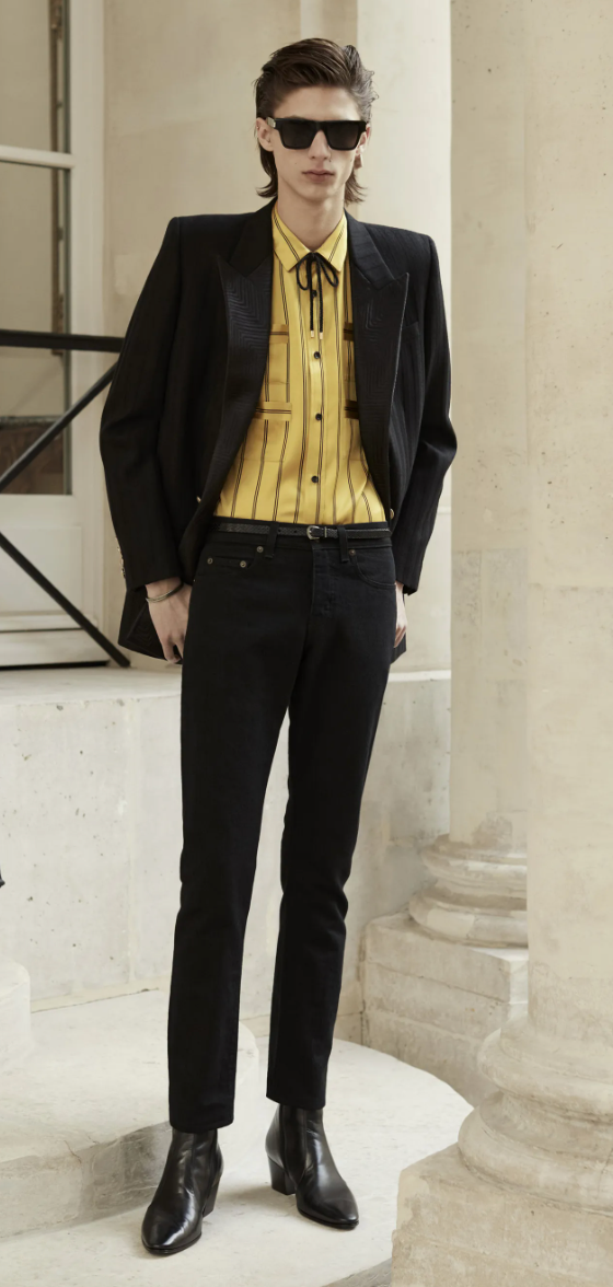A slim man wearing a Yves St Laurent yellow striped button up with a bolo tie, black blazer, pants and shoes.