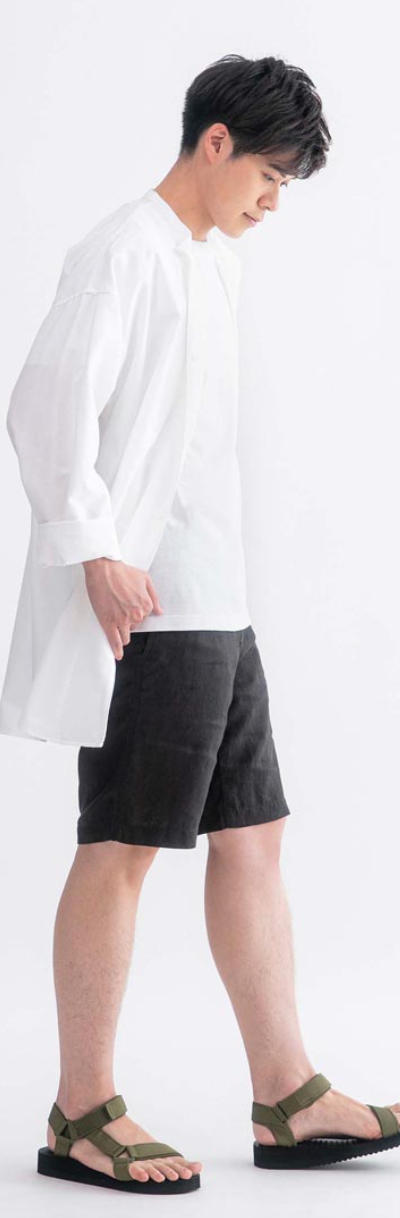 A man wearing a long white Muji button up along with black shorts and olive sandals