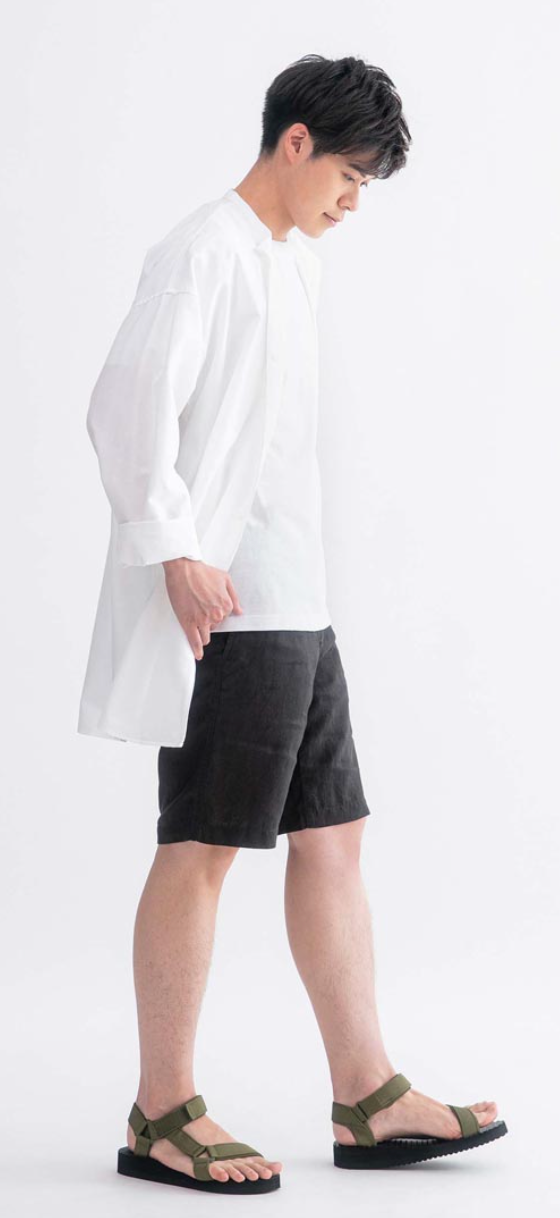 A man wearing a long white Muji button up along with black shorts and olive sandals