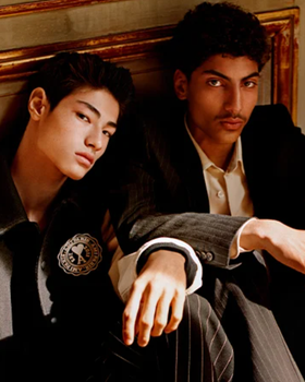 Two men sitting together wear Ami outfits featuring a varsity jacket and suit
