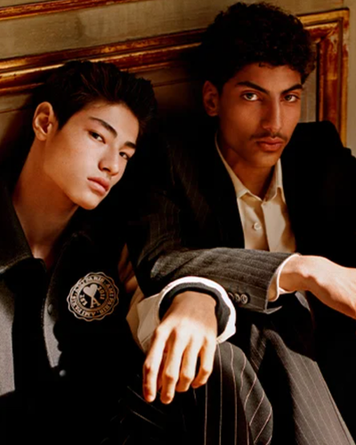 Two men sitting together wear Ami outfits featuring a varsity jacket and suit