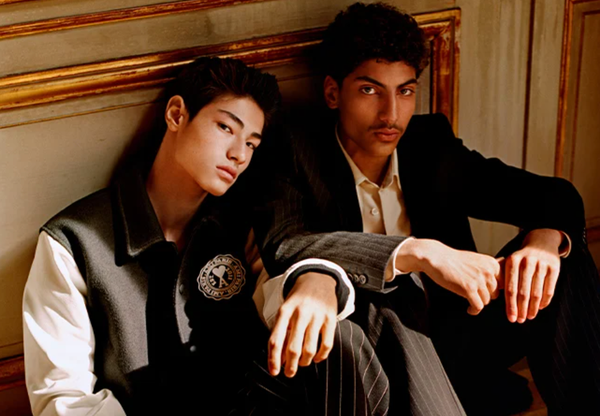 Two men sitting together wear Ami outfits featuring a varsity jacket and suit