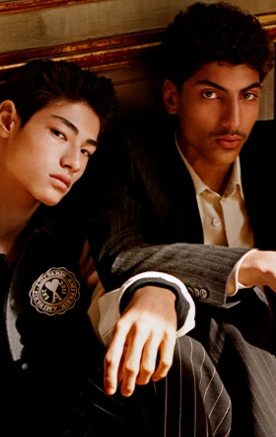 Two men sitting together wear Ami outfits featuring a varsity jacket and suit