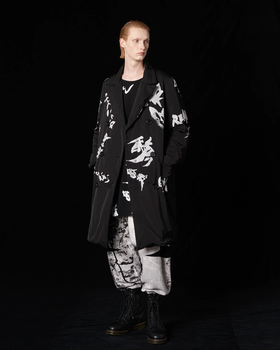 A man with long slicked back hair wearing a Yohji Yamamoto black trenchcoat with white brush strokes across it and black boot