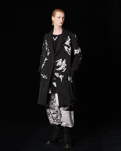 A man with long slicked back hair wearing a Yohji Yamamoto black trenchcoat with white brush strokes across it and black boot