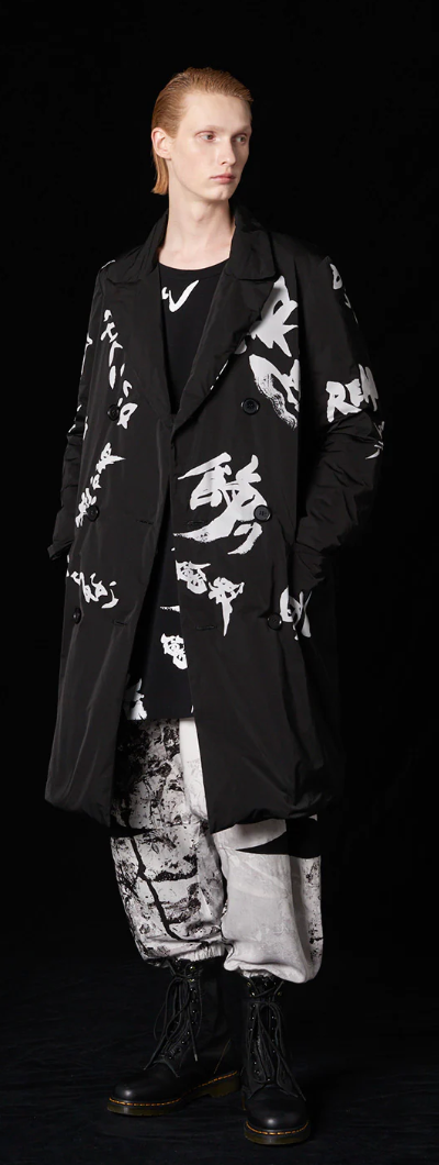 A man with long slicked back hair wearing a Yohji Yamamoto black trenchcoat with white brush strokes across it and black boot