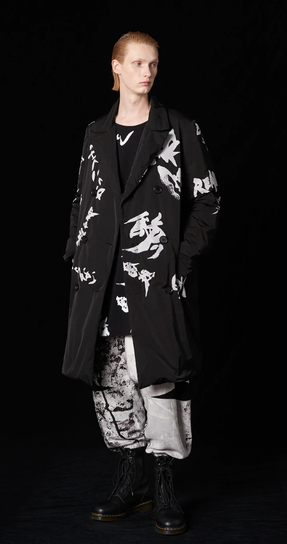 A man with long slicked back hair wearing a Yohji Yamamoto black trenchcoat with white brush strokes across it and black boot