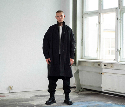 A man wearing a Julius outfit feating a black trenchcoat, black pants and boots along with a white shirt
