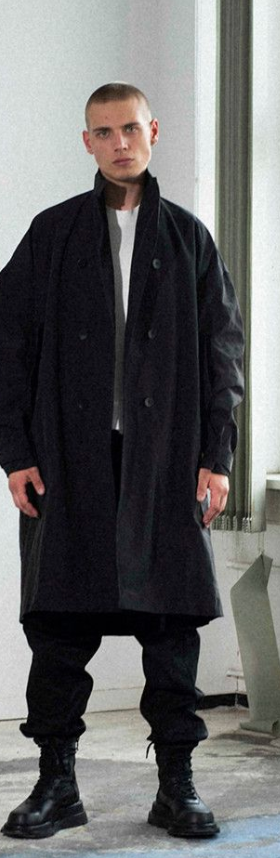 A man wearing a Julius outfit feating a black trenchcoat, black pants and boots along with a white shirt