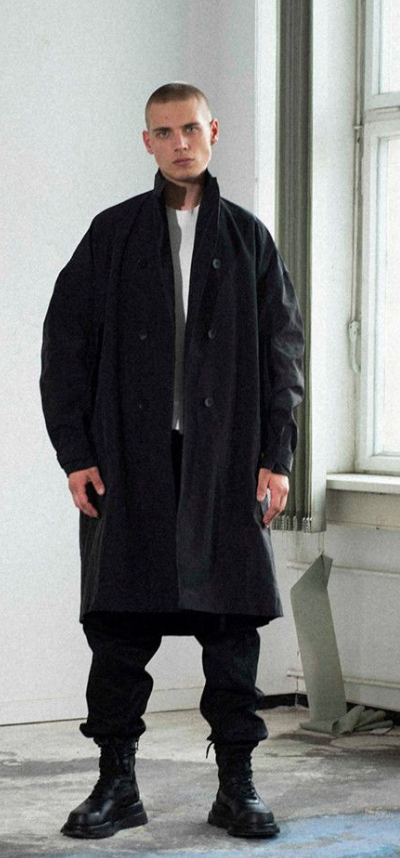 A man wearing a Julius outfit feating a black trenchcoat, black pants and boots along with a white shirt