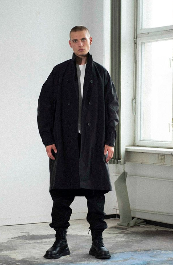 A man wearing a Julius outfit feating a black trenchcoat, black pants and boots along with a white shirt
