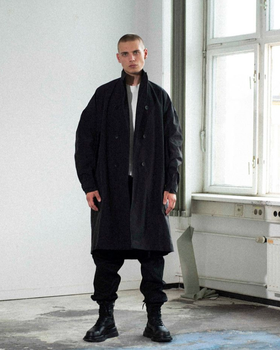 A man wearing a Julius outfit feating a black trenchcoat, black pants and boots along with a white shirt