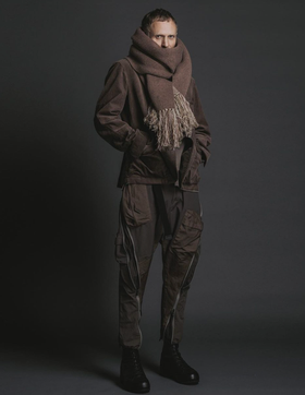 A man wearing The Viridi-Anne oversize brown scarf, a brown jacket with technical looking pants and black hi-top sneakers