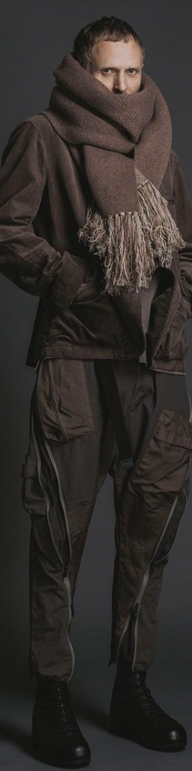 A man wearing The Viridi-Anne oversize brown scarf, a brown jacket with technical looking pants and black hi-top sneakers