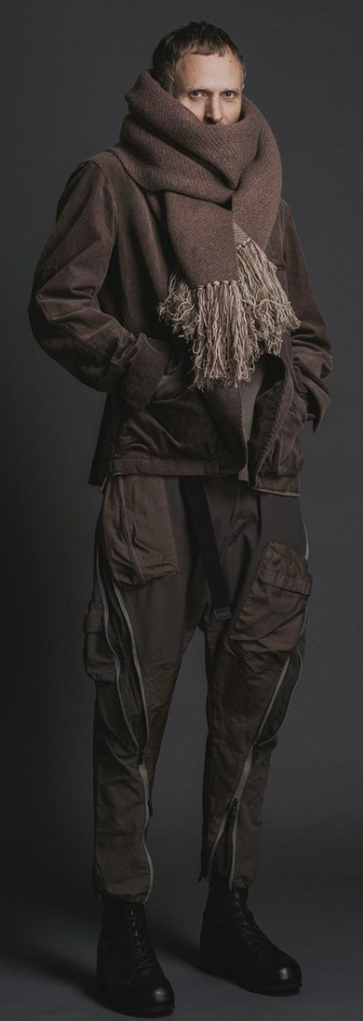 A man wearing The Viridi-Anne oversize brown scarf, a brown jacket with technical looking pants and black hi-top sneakers