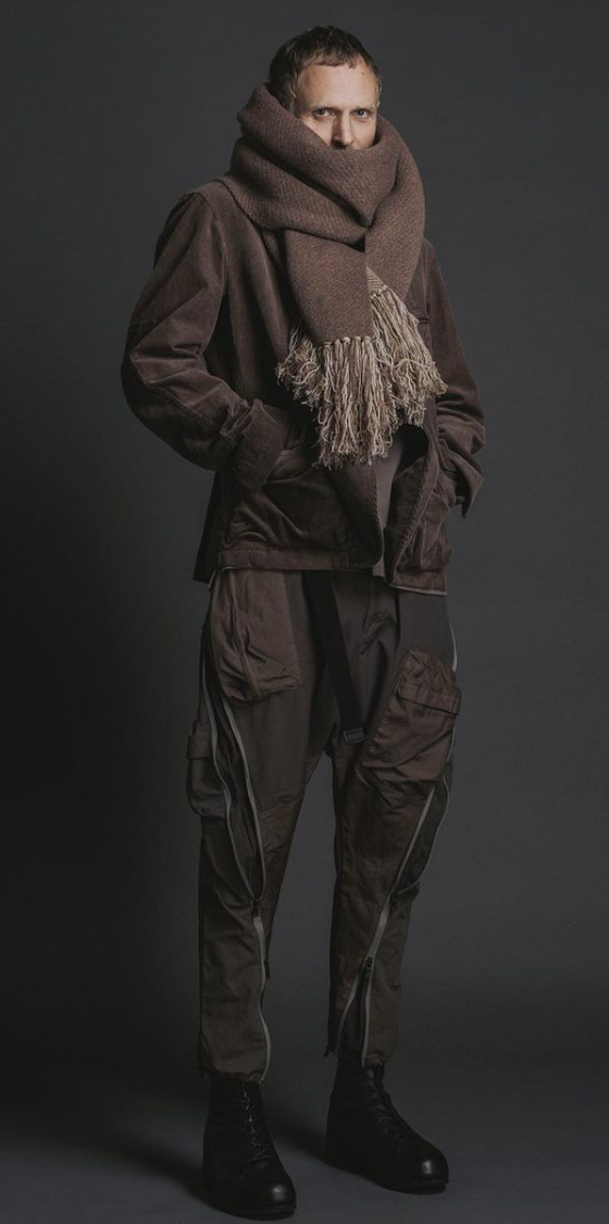 A man wearing The Viridi-Anne oversize brown scarf, a brown jacket with technical looking pants and black hi-top sneakers