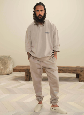 A man wearing Fear of God Essentials loungewear including a beige long-sleeve, matching sweatpants and cream slippers