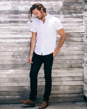 A man wearing a Buck Mason white button-up short-sleeve with black pants and brown loafers