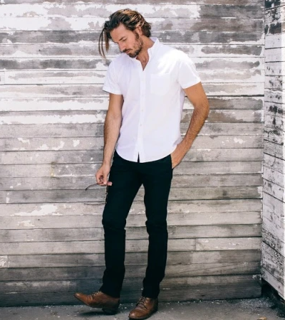 A man wearing a Buck Mason white button-up short-sleeve with black pants and brown loafers