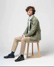 A man sitting and wearing an ISTO green jacket, beige pants and black shoes