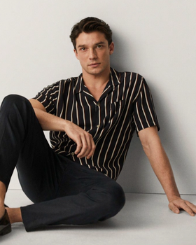 A man sitting against a wall wearing a Mango striped short-sleeve black button up and black tapered pants