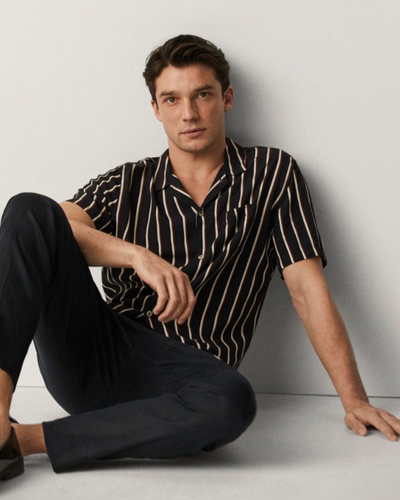 A man sitting against a wall wearing a Mango striped short-sleeve black button up and black tapered pants