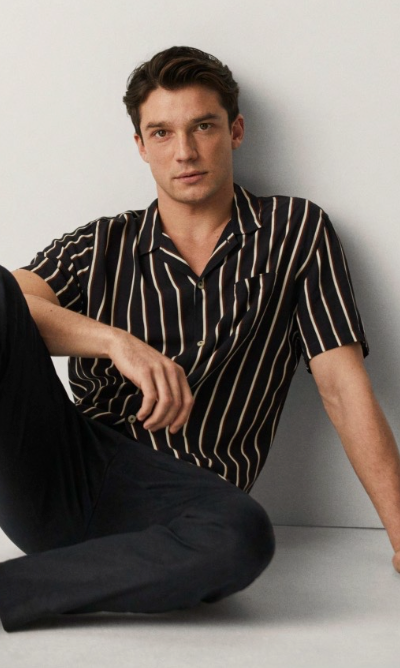A man sitting against a wall wearing a Mango striped short-sleeve black button up and black tapered pants