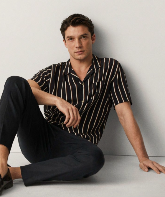 A man sitting against a wall wearing a Mango striped short-sleeve black button up and black tapered pants