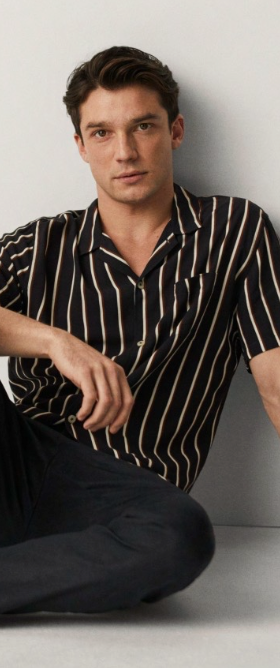 A man sitting against a wall wearing a Mango striped short-sleeve black button up and black tapered pants