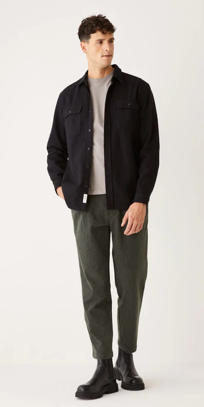 A man wearing a black Frank & Oak button up shirt with dark green pants and black boots