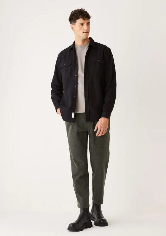 A man wearing a black Frank & Oak button up shirt with dark green pants and black boots