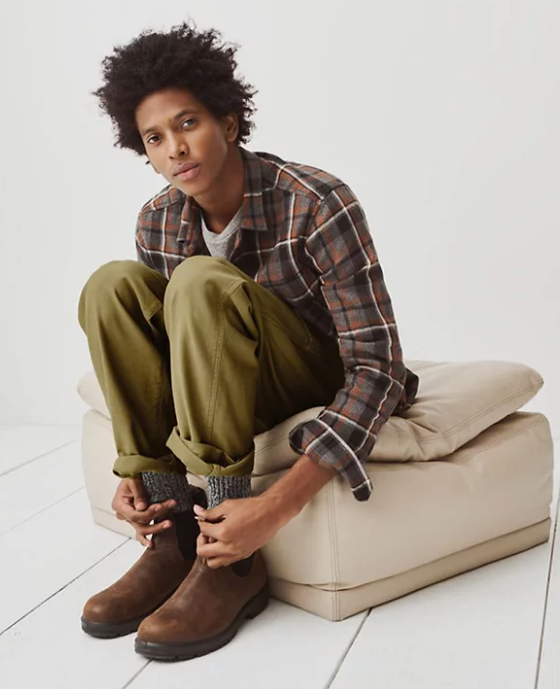 A man sitting and wearing a Madewell plaid button up, green pants and brown blundstone boots