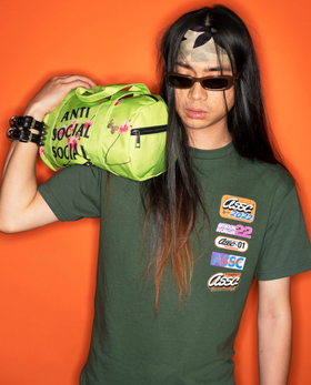 A man with long hair wearing a green Anti Social Social Club t-shirt while holding a lime green small duffle bag