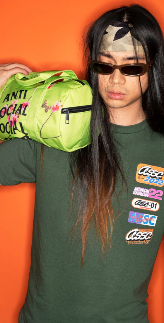 A man with long hair wearing a green Anti Social Social Club t-shirt while holding a lime green small duffle bag