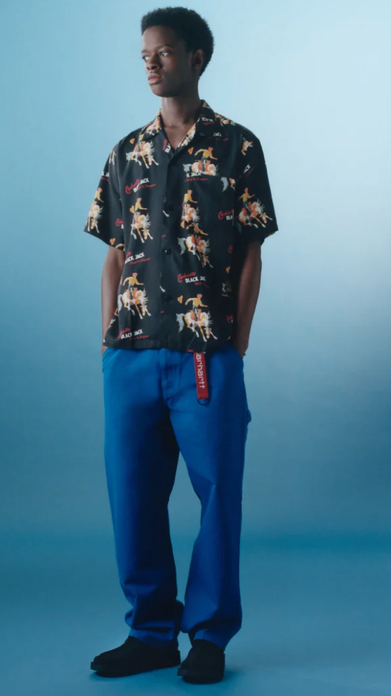 A man wearing a Carhartt WIP black graphic button-up with blue pants and branded carhartt belt