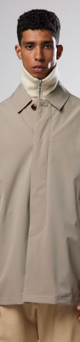 A man wearing a beige NN07 trench-coat with a beige turtle neck and brown pants