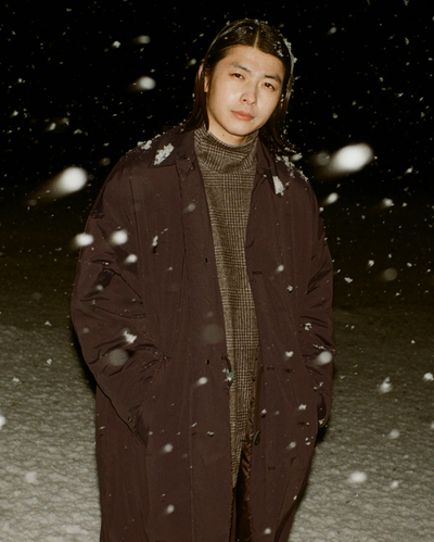 A man wearing a brown N. Hoolywood overcoat with a checkered plaid turtle neck