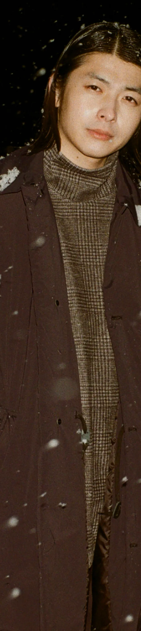 A man wearing a brown N. Hoolywood overcoat with a checkered plaid turtle neck